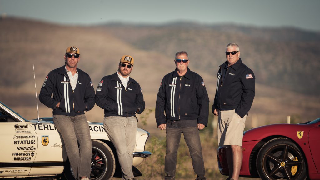 a group of men posing by sports cars on a roadside