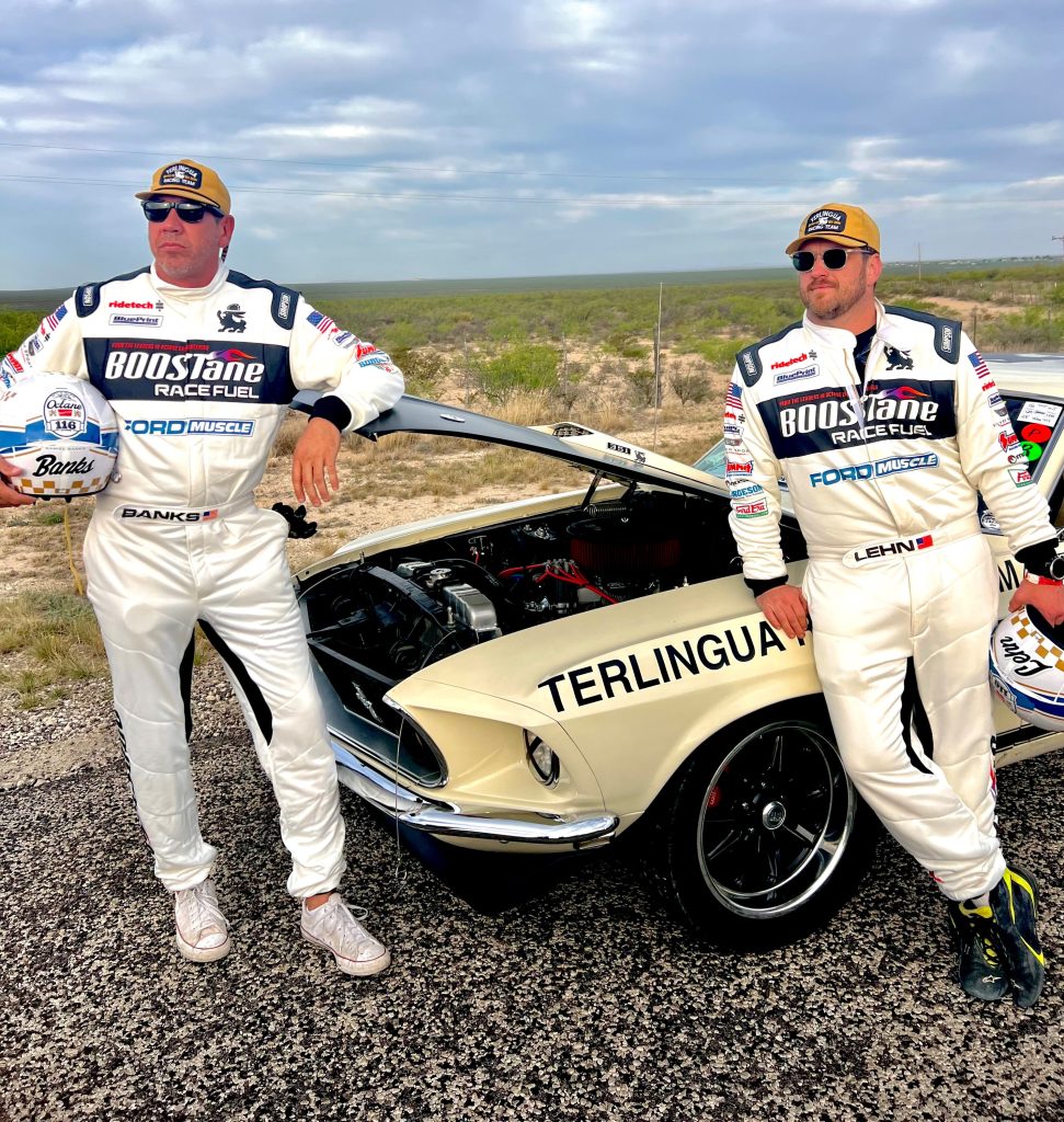 two men in race suits posing next to race car with hood open